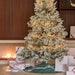 A flocked Christmas tree with warm white string lights stands in a decorated indoor setting, surrounded by wrapped gift boxes and a green tree skirt. A fireplace is visible in the background against a dark wood wall.