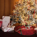 A flocked Christmas tree decorated with warm white lights, gold ornaments, and red ribbons, with a red patterned tree skirt. Several white gift boxes tied with red ribbons are stacked on the left against a wood-paneled wall.