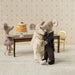 Two toy mice in wedding attire standing next to a cake on a small table with decorative wallpaper in the background.