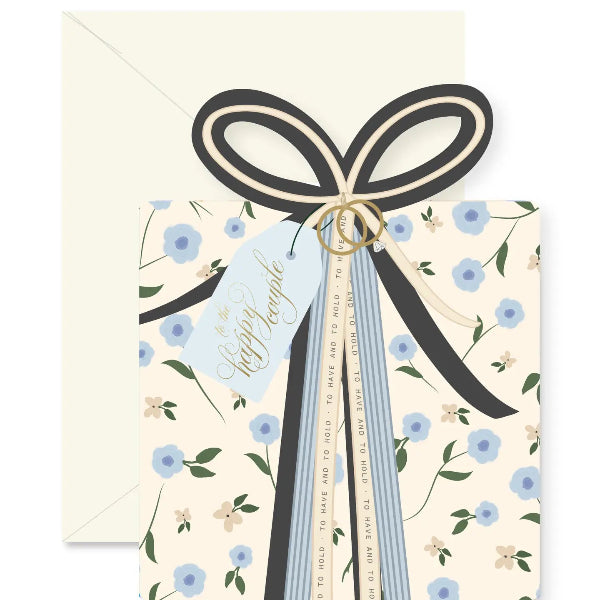 A floral gift tag or card with a cream envelope behind it. The tag has a cream background with small blue flowers, green stems, and a black ribbon bow tied around it, with a light blue hanging tag reading 'to the happy couple'.