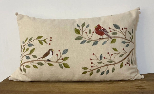 A square beige linen throw pillow with embroidered birds and leafy branches, resting on a wooden surface against a plain white wall.