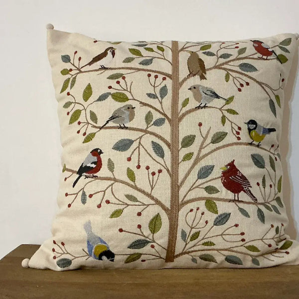A square decorative throw pillow with a detailed embroidered design of a tree with birds and berries, resting on a wooden surface against a plain white wall.
