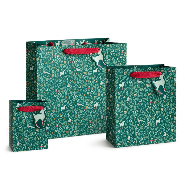 Three green gift bags with a festive Christmas pattern of white deer, red berries, and small ornaments, featuring red paper handles, arranged on a white background.