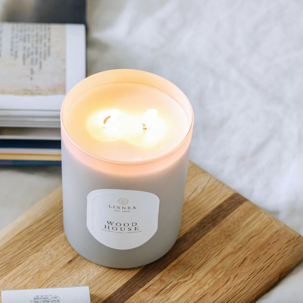 A lit double-wick candle in a frosted white glass jar sits on a wooden board with a soft neutral background and an open book nearby. The label reads LINNEA EST.
