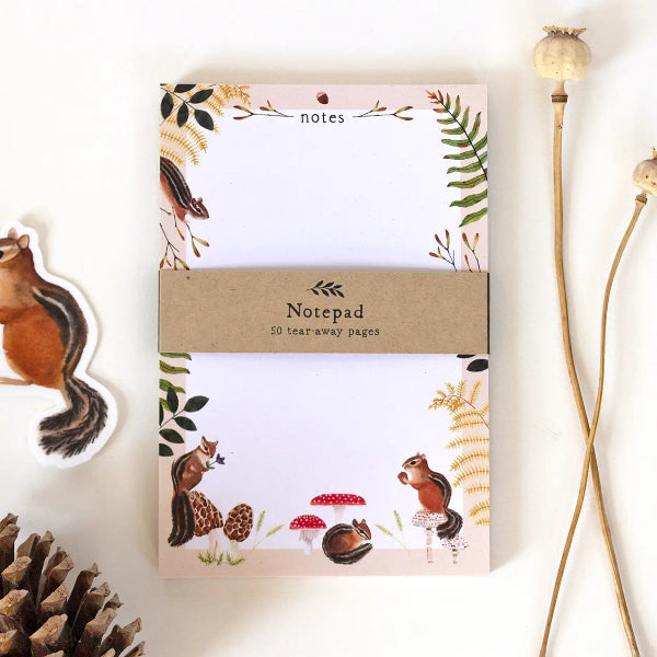 A forest-themed notepad with cute squirrel and mushroom illustrations, wrapped in a kraft paper band labeled 'Notepad 50 tear-away pages'. The notepad is displayed in a styled flat-lay with pinecones and dried poppy stems on a white background.